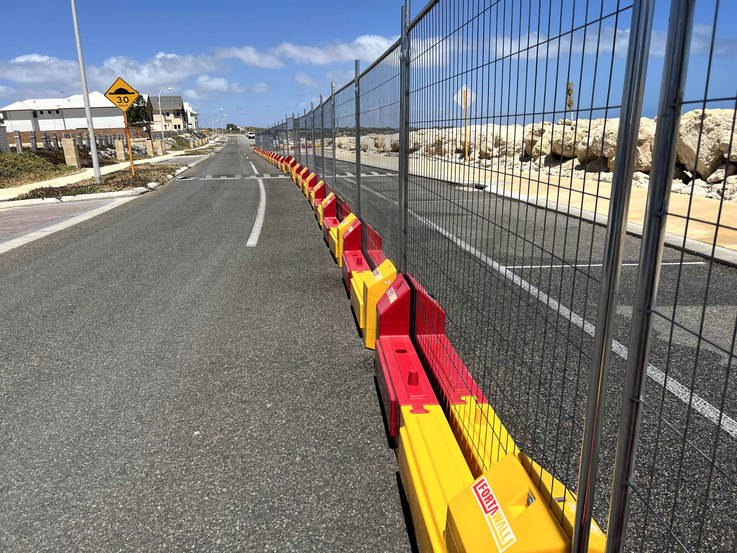 Gallery: A Forta Wall barrier system with yellow and red bases lines a road. A wire mesh fence sits atop the barriers, with a speed bump sign visible.
