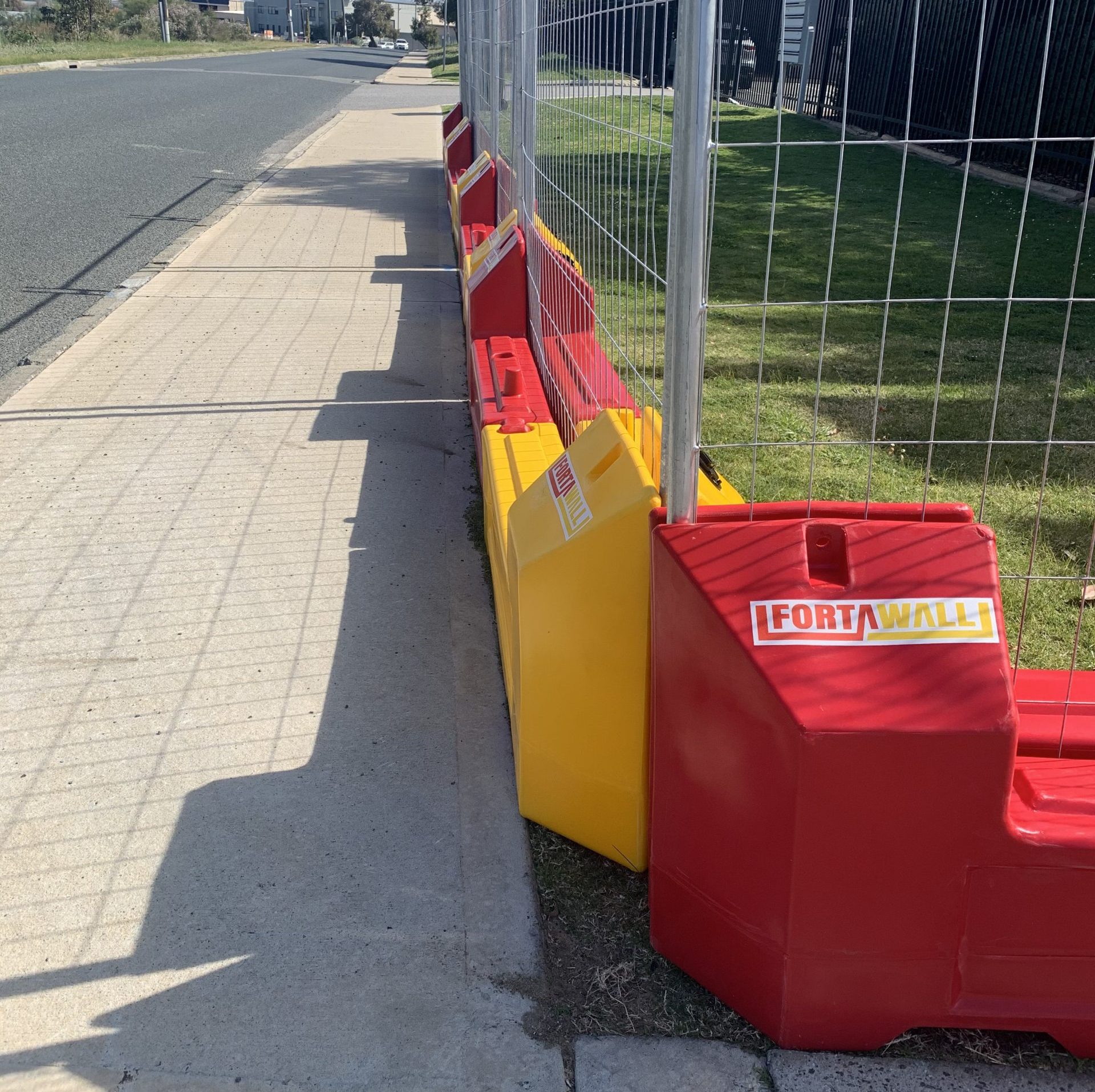 Red and yellow FortaWall barriers with wire mesh fencing along a sidewalk. Construction site safety equipment for home projects.